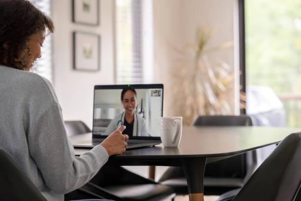 A female patent of African decent meets with her doctor remotely via a video call. She is sitting at her kitchen table with her laptop out and her prescription on the table with her. The doctor can be seen on the screen wearing a white lab coat as she talks with her patient.